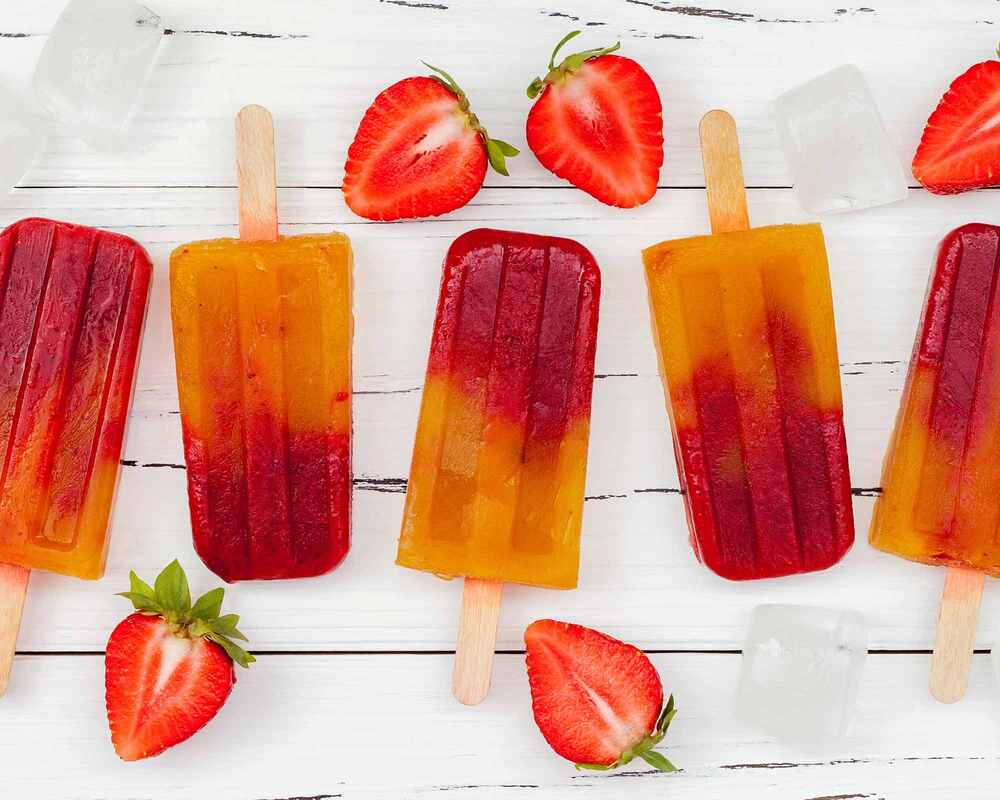 Alcoholic Icy Poles that are a mix of red and orange in colour. They are on a table and surrounded by ice and strawberries