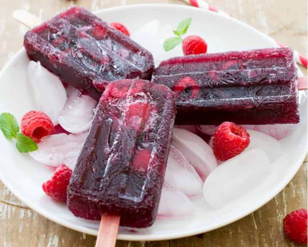 Three raspberry sangria popsicles on a white plate. They are surrounded by raspberries and ice blocks